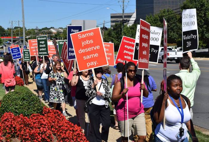 A multiracial group of protesters, many of them using guide canes, holding large and colorful printed posters as they march outside beside a bed of red flowers in the sunlight. The messages say,  - Do you support equality or exploitation?  - Minimum wage should protect everyone.  - Same jobs deserve same pay.  - SourceAmerica exploits people with disabilities.  - CEO Salary: $500,000. Worker Pay: 20 cents.