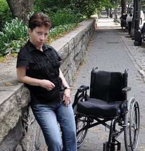 White woman standing and leaning against a stone wall lined against city street. She has short cropped brown hair and is wearing a short-sleeved black shirt and blue denim jeans. Next to her is a manual chair with black cushion and back.
