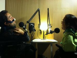 Image of two people in a recording booth with microphones angled near their faces. On the left is a Latino man in a wheelchair using a nasal mask for respiratory support. On the right is an Asian woman in a wheelchair with glasses. They are in conversation with one another.