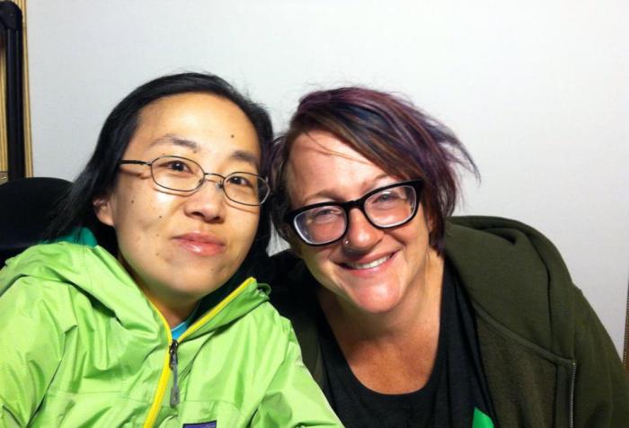 Two people smiling at the camera sitting side-by-side. On the left is an Asian woman wearing a green jacket and glasses. On the right is a white woman with brown hair with purple highlights and glasses.
