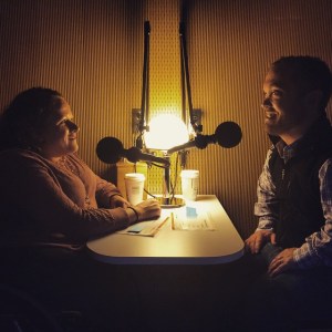 A room with dimmed lighting, two people are sitting across from one another with microphones in front of them. On the left is a middle-aged white woman in a wheelchair with long brown hair. On her right, facing her, is a middle-aged white man with short brown hair.