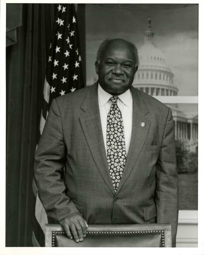Congressman Major Owens, U.S. House of Representatives, 1983-2006. Major Owens Collection, Brooklyn Public Library – Center for Brooklyn History. Photo of an older Black man who is wearing a suit and tie standing with one hand placed on the back of a chair. Behind him is the American flag on a stand to the left of him and a backdrop showing the US Capitol building. He is smiling at the camera.