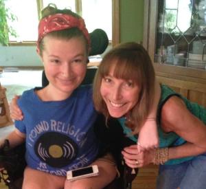 Photo of a young white woman in a wheelchair. Her hair is tied up with a red bandana. She has a blue t-shirt on, and an iPhone is on top of her left lap. She has her arm around a middle aged white woman who has shoulder-length blonde brown hair. She is wearing teal tank top and blue jeans. Both are smiling at the camera.