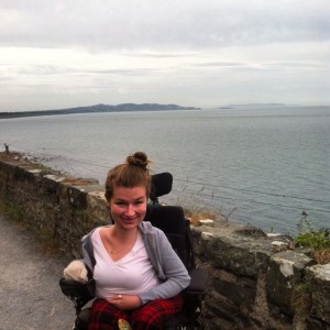 Image of a young white woman with her blonde hair tied up in a bun. She is outdoors behind a body of water and a wall made of stone. She is wearing a v-neck white t-shirt with a gray cardigan sweater. She is smiling at the camera.