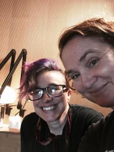 Image of two white women in a recording studio. The one in the center has a mohawk w/ pink highlights. She is wearing glasses. The woman on the right side has short red hair and she is smiling.