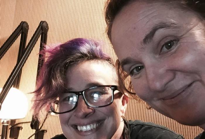 Image of two white women in a recording studio. The one in the center has a mohawk w/ pink highlights. She is wearing glasses. The woman on the right side has short red hair and she is smiling.