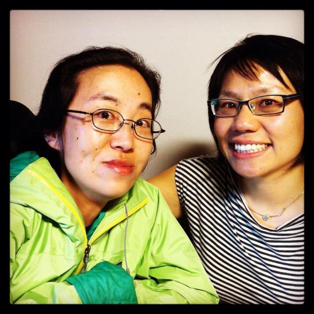 Two Asian American sitting side-by-side. The woman on the left is in a wheelchair; she is wearing a green rain jacket and glasses. The woman on the right has short black hair, glasses and is wearing a striped-t-shirt. They are both smiling at the camera.