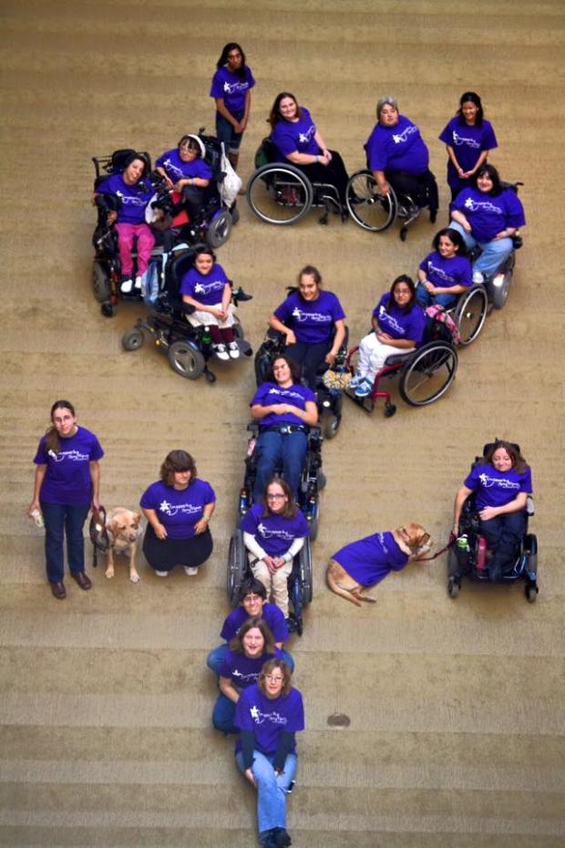 Photo from an overhead perspective of a bunch of young women with disabilities,many in wheelchairs, all wearing purple t-shirts, they formed a female symbol
