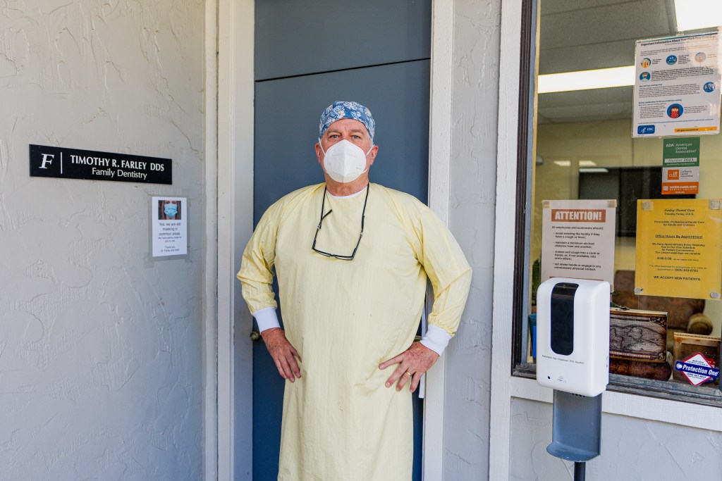 Dr. Timothy R. Farley, DDS stands in front of the entrance to his practice. A sign to his left reads “F | Timothy R. Farley DDS. Family Dentistry.” Below this sign is another with a picture of a person wearing a surgical mask that reads “Yes, we are still masking in common areas. We have plenty inside for you to use. Thank you: Dr. Farley and Staff.” Photo by Brian Ringo, 2023.