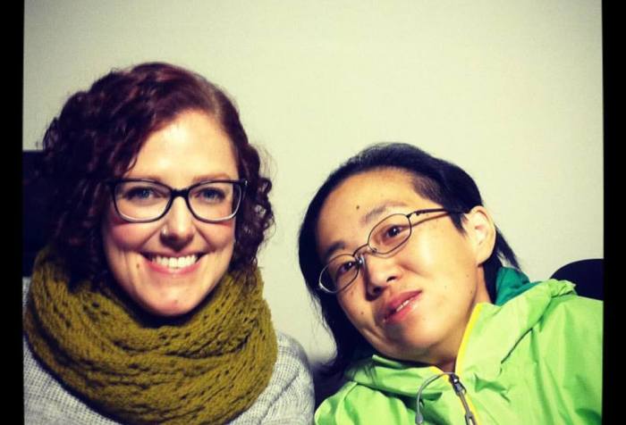 two women in wheelchairs sitting side-by-side against a plain white background. The white woman on the left has curly red hair and is wearing glasses, a gray sweater an a green scarf wrapped around her neck. The Asian American woman on the right has her head tilted toward her friend, she has black hair and is wearing glasses and a green jacket.