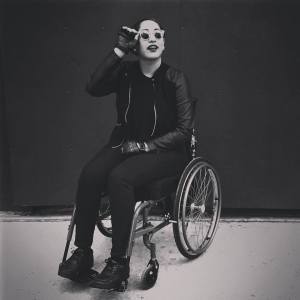 Image Description: black and white photo of Annie, a tall, light brown skinned person with slicked-back hair, dressed in all black and sunglasses, seated in a manual wheelchair.