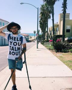 Young man with one above-the-knee amputation on his left leg. He is balancing two crutches with his left arm. He is wearing denim shorts, that, glasses and a beard. Behind him is a neighborhood by the beach with a row of palm trees.