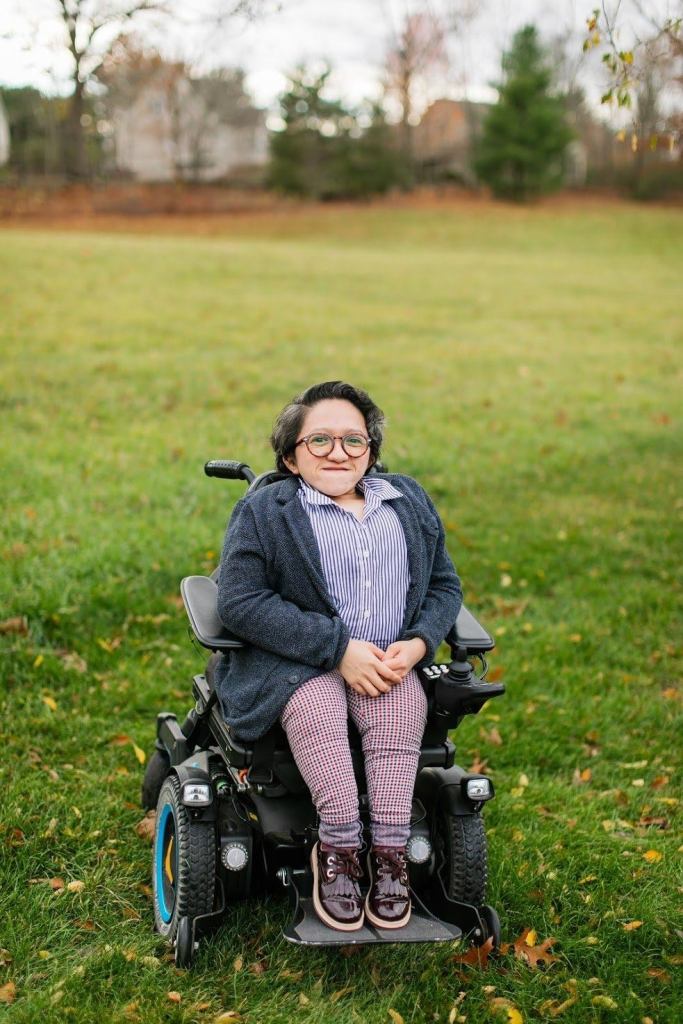 This is a photo of Sandy Ho sitting in her power wheelchair in the middle of a field of green grass, with houses blurred in the background. She has short dark wavy hair and tortoise shell glasses on. Sandy is wearing maroon shoes, plaid pants, a dark gray cardigan, and a button-up blue and white striped collar shirt. She is smiling at the camera with her hands in her lap.