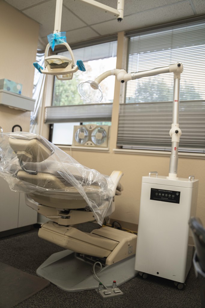 Patient chair covered in plastic, which is replaced after every patient. Above it hangs the exam room light, where the touched surfaces are covered in plastic, which is also replaced after every patient. In the background is the intake/exhaust fan that vacuums and ventilates the room. On the right of the chair is the suction air purifier which runs during aerosol producing procedures, removing aerosols from the air and running them through an air purifier with a UV-c light that neutralizes viruses and bacteria. Photo by Brian Ringo, 2023.