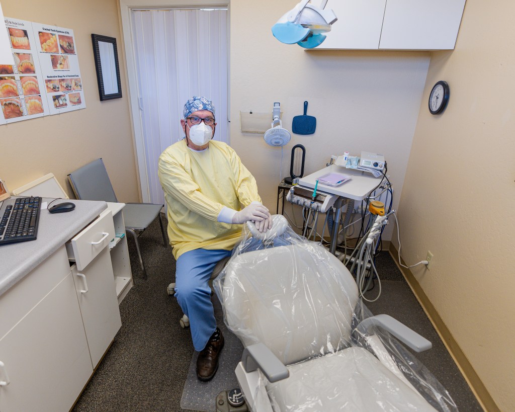 Dr. Timothy R. Farley, DDS sits behind an empty chair in an examination room in his dental practice. He is wearing a KN95 mask and other personal protective equipment (PPE). The dental chair in front of him is covered in plastic that is changed after every patient. Behind him is the flexible door that, along with the exhaust fan and air purifiers, helps mitigate the amount of aerosol generated through dental procedures from potentially contaminating common areas.