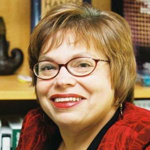 Judy Heumann with short brown hair and red glasses looks directly at the camera smiling, wearing a red blouse.