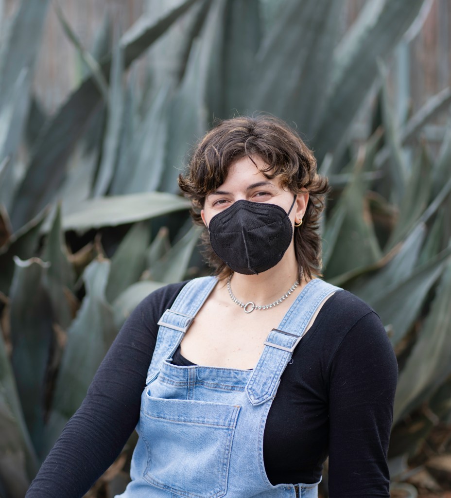 Cecilia Senocak sitting and “smizing” at the camera with their eyes over their black KN95 mask. Behind them is a cluster of agaves. Photo by Brian Ringo, 2023.
