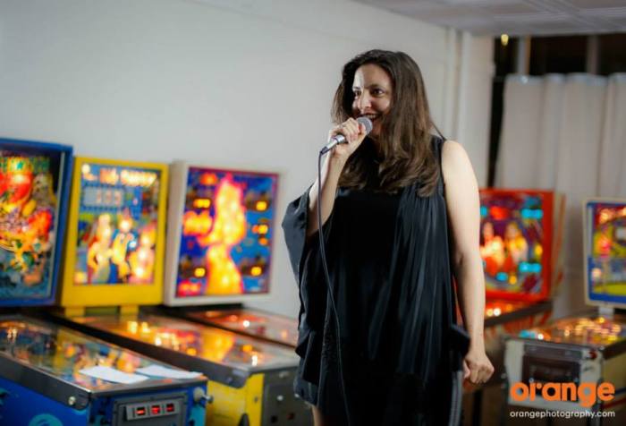 Photo of a woman with long brown hair holding a microphone with pinball machines in the background.