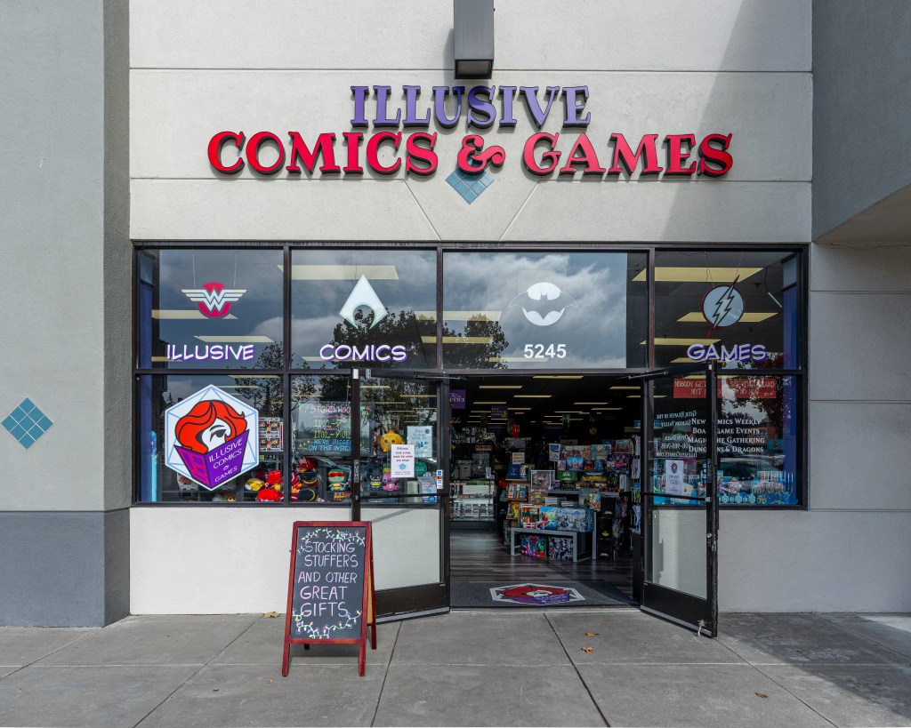 The storefront of Illusive Comics and Games. Beneath the business title are many windows with comic book icons like Wonder Woman and Batman, among others. The business’ logo is a red-haired person reading a purple comic book. The doors of the business are wide open, a sign on the door encourages mask use, and a chalk sign out front reads “Stocking stuffers and other great gifts” with little holiday lights drawn around the text. Photo by Brian Ringo, 2023.