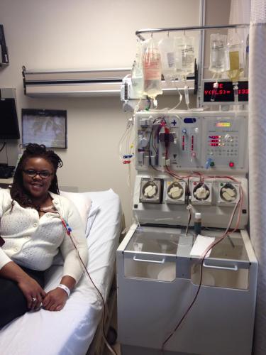 A Black woman on a hospital bed and she is receiving a blood transfusion. There is a tube with blood connected to a large machine with hanging bags of blood.
