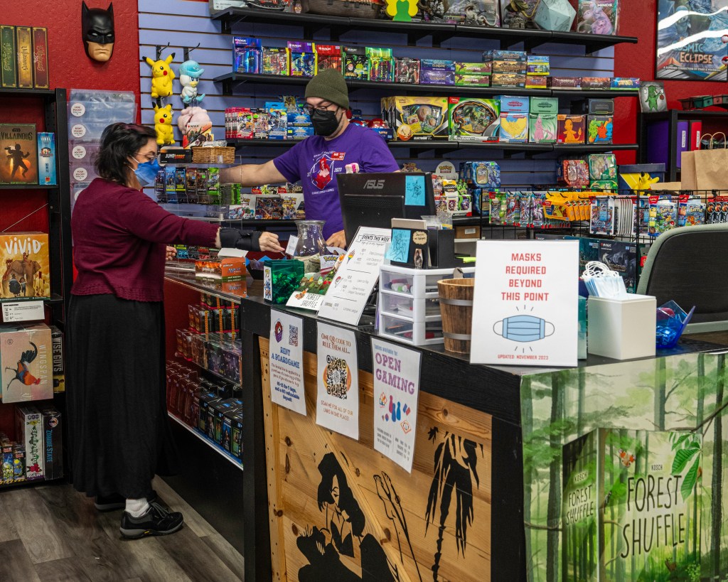 Illusive Comics and Games LLC owner Anna Warren wears a blue ombre KN95 style mask across the cash register from employee Will, who is wearing a black KN95 style mask. Brightly colored games, Pokémon merchandise, and other goodies stuff every flat surface and wall. A sign reading “Masks required beyond this point: Updated November 2023” sits on the corner of the cash register area with a container of free surgical masks next to it. Photo by Brian Ringo, 2023.