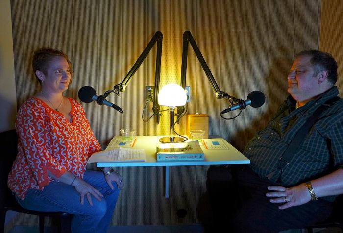 Image of two people facing each other in a recording booth with microphones in front of them. On the left is a middle-aged white woman with short red hair. She is wearing red-print shirt and blue jeans. On the right is an older white man with short brown hair. He is wearing a dark shirt with suspenders.