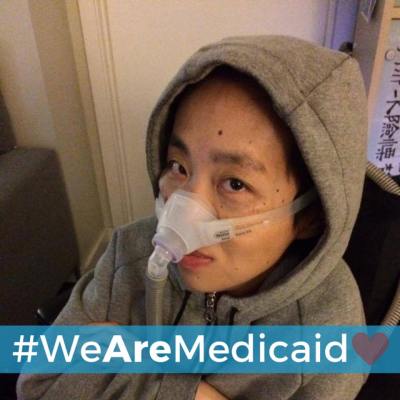 Image description: Asian American woman wearing a gray hoodie with her hands crossed across her chest. She is sitting in a wheelchair and had a BiPap mask over her nose with a tube. Below is a light blue horizontal bar with white text that reads: "#WeAreMedicaid." Next to the text on the right side is a graphic of a pink heart