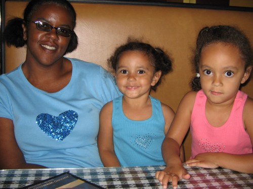 A young Black woman with hair tied in pig tails wearing a blue t-shirt. She is sitting at booth with two of her young biracial daughters, both are wearing tank tops with their hair tied back.