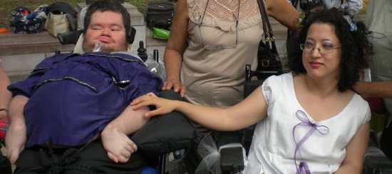 Photo of a man using a ventilator wearing a put;poe t-shirt on the left side. On the right is a woman with dark curly hair wearing glasses and a white dress. She is seated and has her hand touching her husband's arm