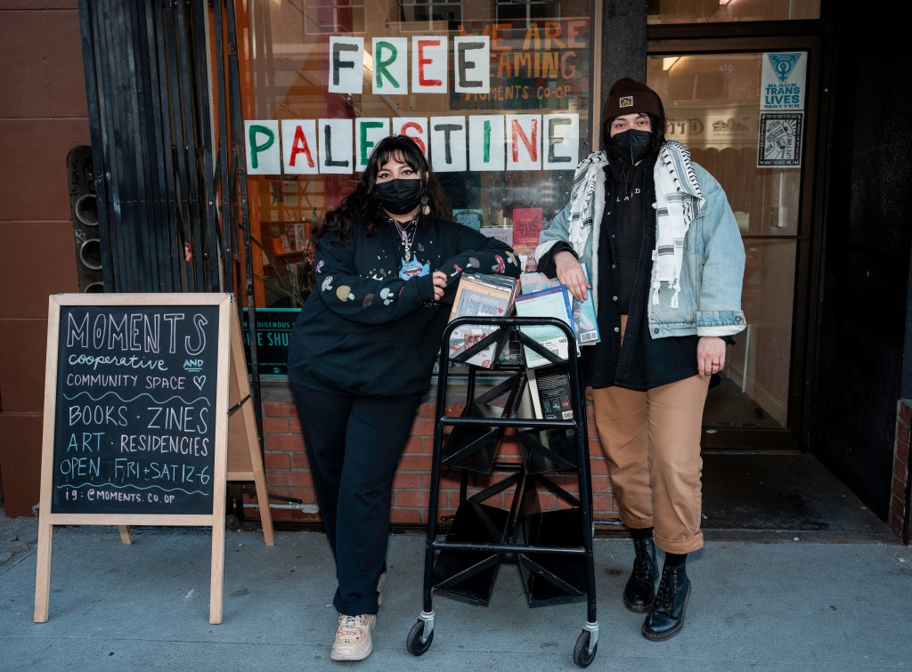 Moments Cooperative and Community Space members Samantha and D posing outside the bookstore entrance wearing black KN95 masks. They are leaning against a cart of books for sale. Next to them is an A-Frame reading "Moments Cooperative and Community Space. Books. Zines. Art. Residencies. Open Fri-Sat 12-6. IG: @Moments.co.op.”Behind them in the window reads "FREE PALESTINE.” Photo by Brian Ringo, 2023.