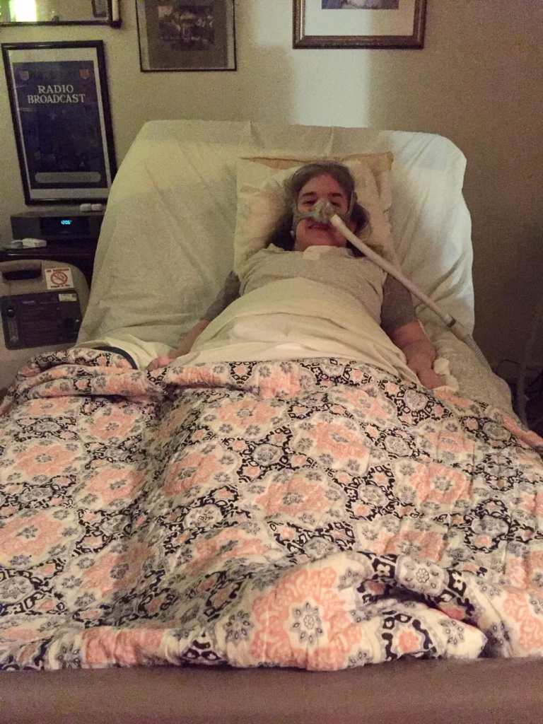 First day home from the hospital. Ingrid Tischer, middle-aged white woman wearing a Bi-Pap mask in a hospital bed under a colorful quilt.