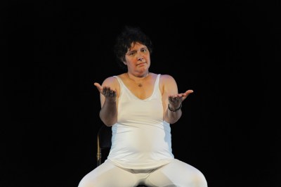 Photo of a person dressed in a white tank top and white pants. The person's curly dark hair and the person's palms are open and arms outstretched. The background is completely dark.
