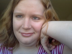 Young white woman with long reddish-blond hair. Her left arm is raised and her hand bent against her face. She is smiling.