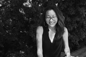Black and white photo featuring a young queer disabled Korean woman. She is wearing glasses and a black v-neck tank top. She is standing in front of a bunch of trees with leaves all around her. 