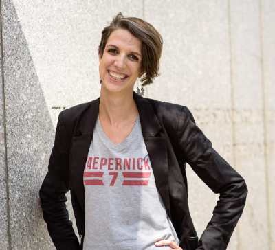 Photo of a white woman, smiling and standing with her hands on her hips and head slightly tilted to the left, in front of a gray granite wall. She has short brown hair in an asymmetrical haircut, longer on her left side and shorter on her right side. She is wearing dangling silver earrings and a gray t-shirt that reads "Kaepernick 7" in red, underneath a black blazer. 