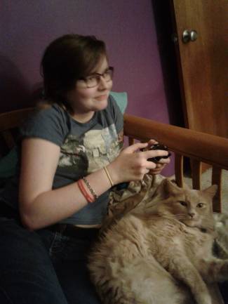 Young white woman with short brown hair and glasses holding a game controller. She is wearing a t-shirt and behind her is a purple wall. A cat is sitting on her lap.