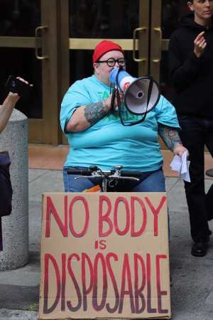Max Airborne, a fat white person wearing blue jeans, an aqua t-shirt, a red knit beanie and round glasses, sits on a scooter in front of San Francisco ICE headquarters, speaking through a bullhorn. Resting on the front of their scooter is a large cardboard sign hand-painted with the words "No Body Is Disposable." On their left someone is interpreting in ASL, and on their right someone is recording audio using a handheld device.