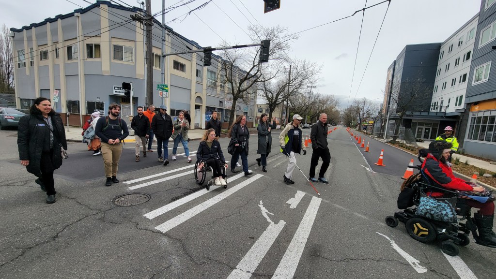 A group of a dozen adults, including some people using white canes, manual and power wheelchairs, cross a sidewalk in Seattle Washington. The sky is grey and everyone is bundled up in warm clothes. There are apartment buildings on either side of the street, and trees without leaves. 