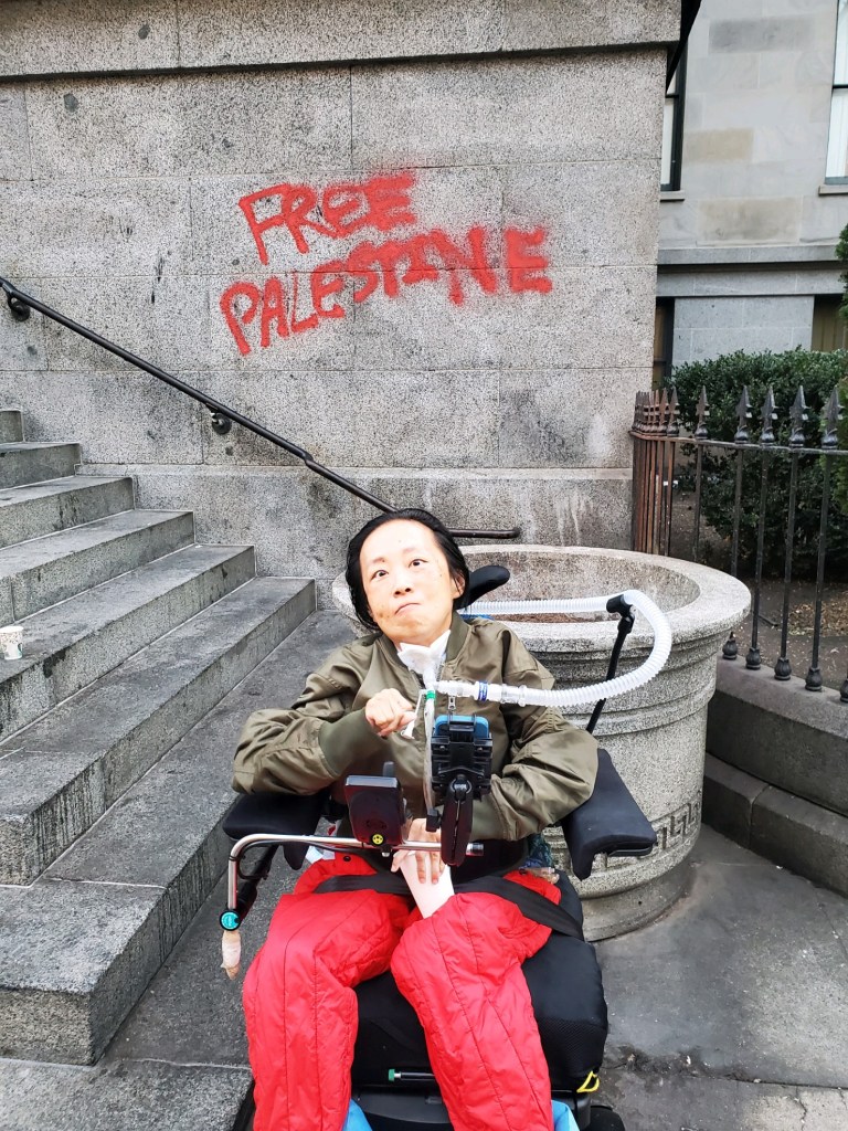 A graphic with a red background and in the center is a photo of an Asian American disabled woman in a wheelchair with a tracheostomy attached to a ventilator. Behind her is a building FREE PALESTINE written in red paint