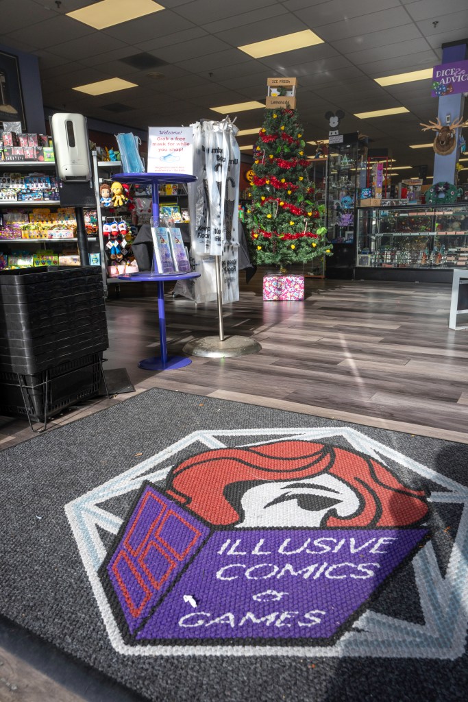 A sign reading “Welcome! Grab a free mask for while you shop. Required in event area and from 11a-12p daily” greets customers upon first entering Illusive Comics. It is nestled between the shopping baskets, hand sanitizer, and umbrella covers. A Christmas tree with gifts underneath it appears in the background along with assorted merchandise. Photo by Brian Ringo, 2023.