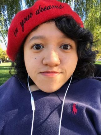 A headshot photo of Sandy Ho, Asian-American disabled woman with dark curly hair sitting in a wheelchair looking into the camera. Sandy is wearing a red beret and the words "In your dreams" are printed on it. She has on a dark blue sweatshirt, and white iphone headphones. The background is sunlit with grass and a willow tree.
