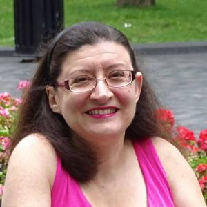 Image description: Caucasian woman with shoulder-length brown hair and glasses is smiling. She is wearing a pink sleeveless dress and is seated in front of a rose bush.