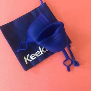 Against a peach colored background, photo of a blue square bag with the word 'Keela' and 2 blue silicone menstrual cups lying on top of the bag.