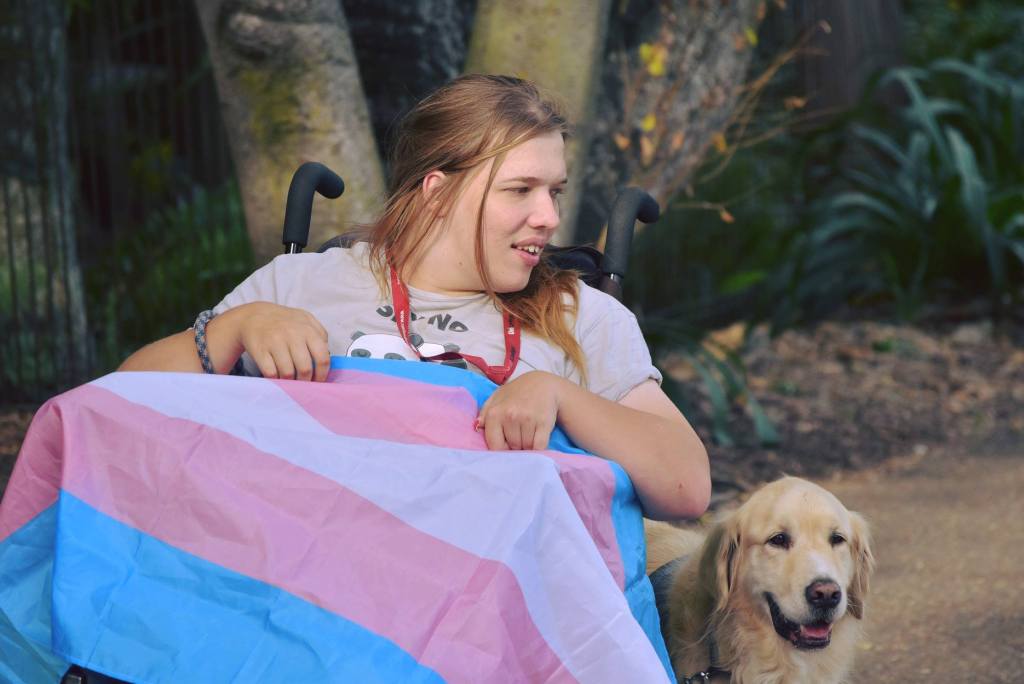 Photo of Annabelle Oxley, a young white woman draped in a trans flag and her assistance dog Jasper, a golden retriever. 