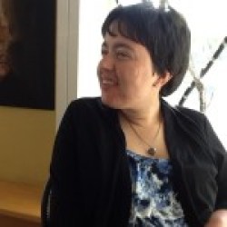 Picture of a white woman seated in a chair. Her head is turned to the right and she's smiling. She has short dark hair and is wearing a floral-print top with a black jacket.