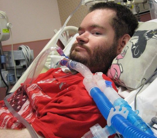 A man with brown hair and a beard sitting upright in a hospital bed. He is using a ventilator.