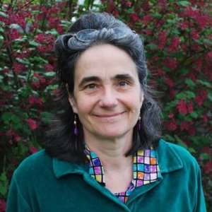 An older white woman with long dark hair. She is wearing a teal shirt and standing in front of a bush with flowers. She is smiling at the camera.