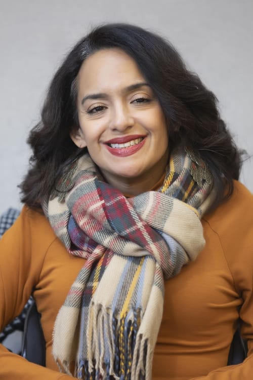 a photo of Ligia Andrade Zúñiga, a woman color with a disability smiling, she has long wavy hair, is wearing a scarf, and has long dangly earrings.