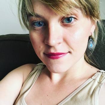 A closeup of a white woman with blonde hair and blue eyes smiling at the camera. She is wearing teardrop earrings.