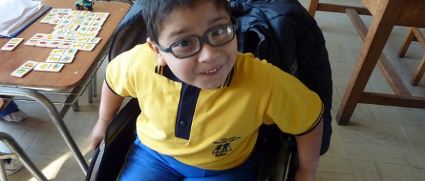 Photo of a young boy in a manual wheelchair. He's wearing glasses and a yellow shirt with blue pants.